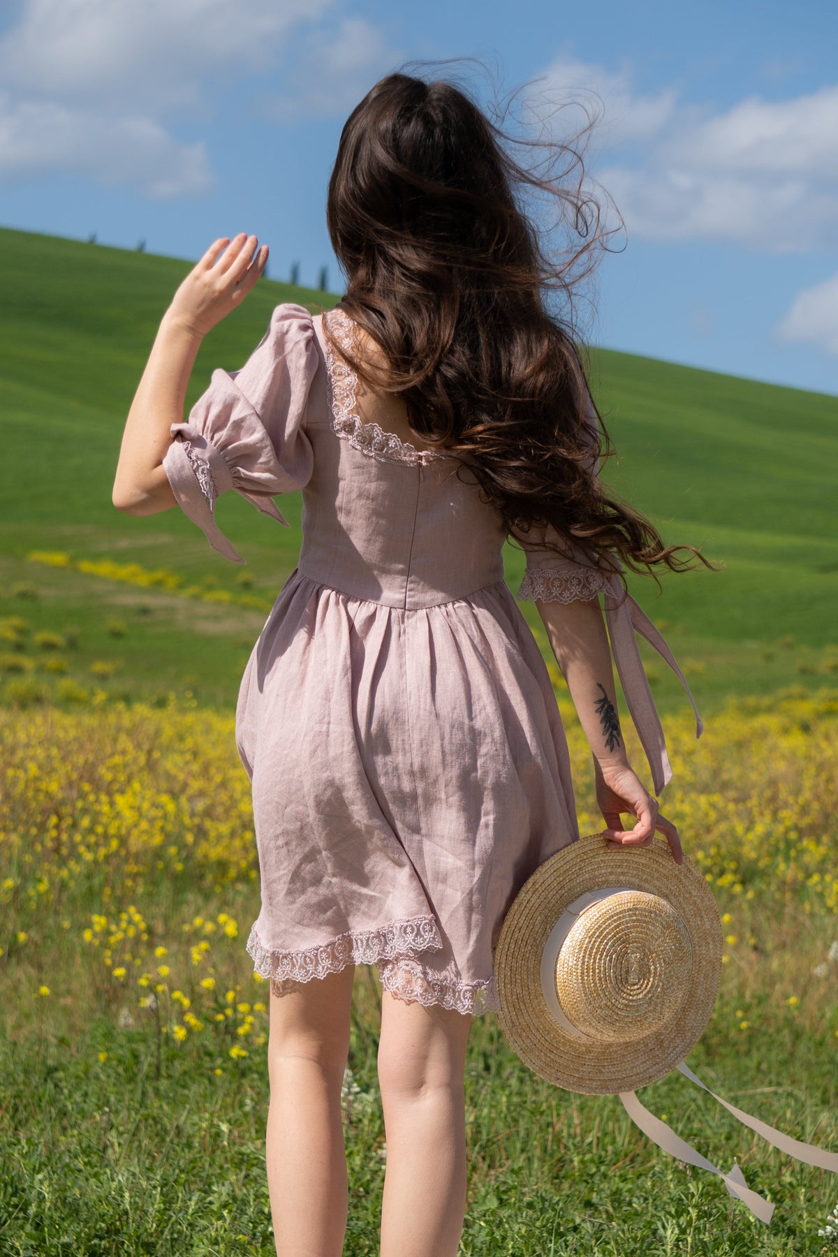 Back view of the April antique pink linen mini dress, holding a straw hat on a green hillside