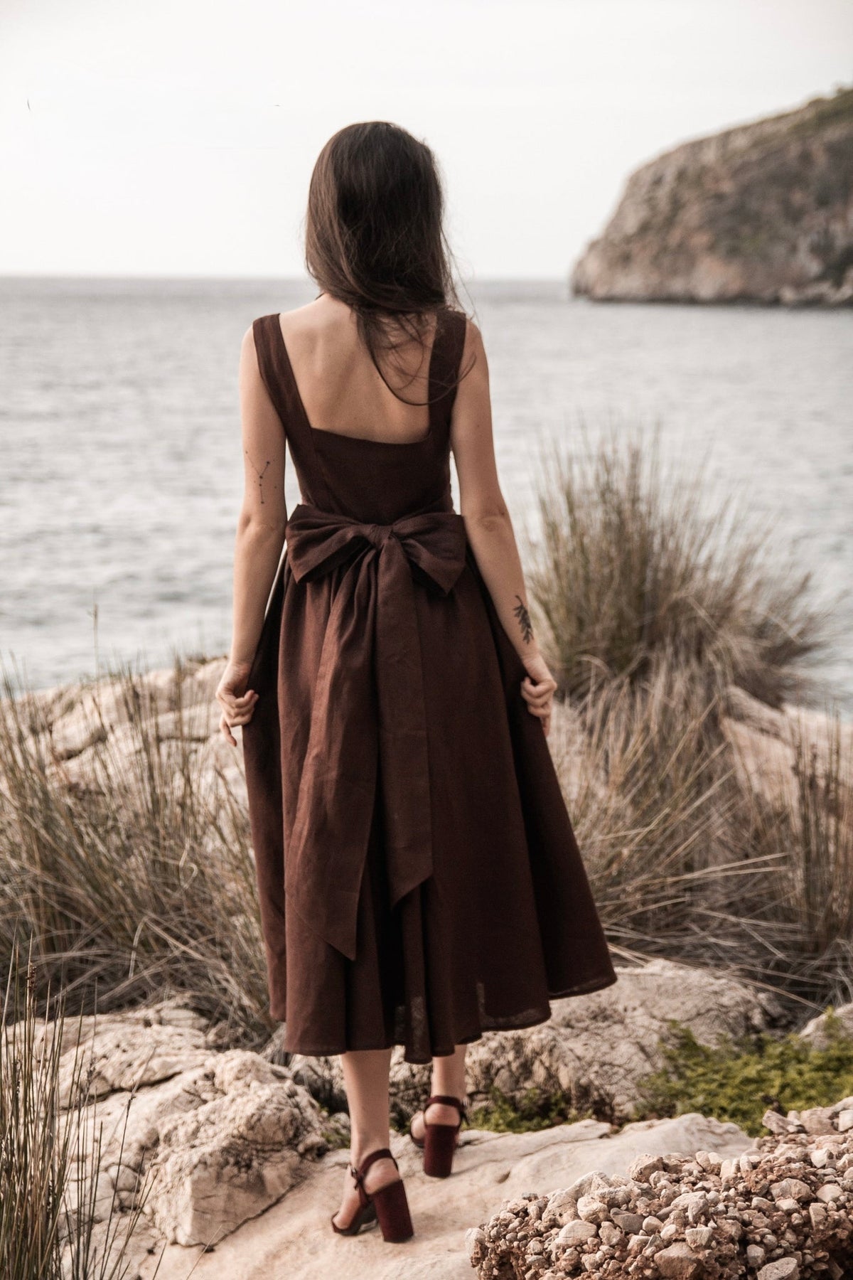 Back view of the Andalusia dark brown linen dress with a large bow at the waist, photographed on a rocky seaside