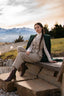 Chic woman seated outdoors in beige wool elegant suite with a green wool coat draped over her shoulders, posing against alpine peaks at sunset; elegant quiet luxury countryside aesthetic.