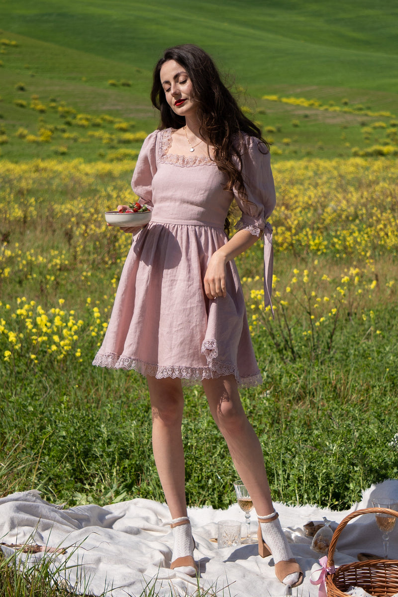 April antique pink linen mini dress with puff sleeves and lace trim, standing on a picnic blanket in a meadow