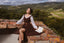 A woman in a brown and white dress sits on a rustic brick ledge overlooking a scenic green valley landscape under a blue sky with clouds, with an open book resting beside her.