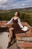 A woman in a brown and white medieval-style dress sits on a rustic brick ledge overlooking a scenic green valley landscape under a cloudy sky, with an open book resting beside her.