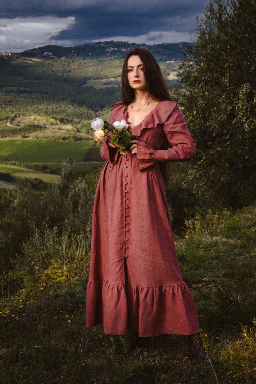 Florence brick red linen maxi dress with a ruffled collar and bell-cuff sleeves, white roses, Tuscan hills under a stormy sky