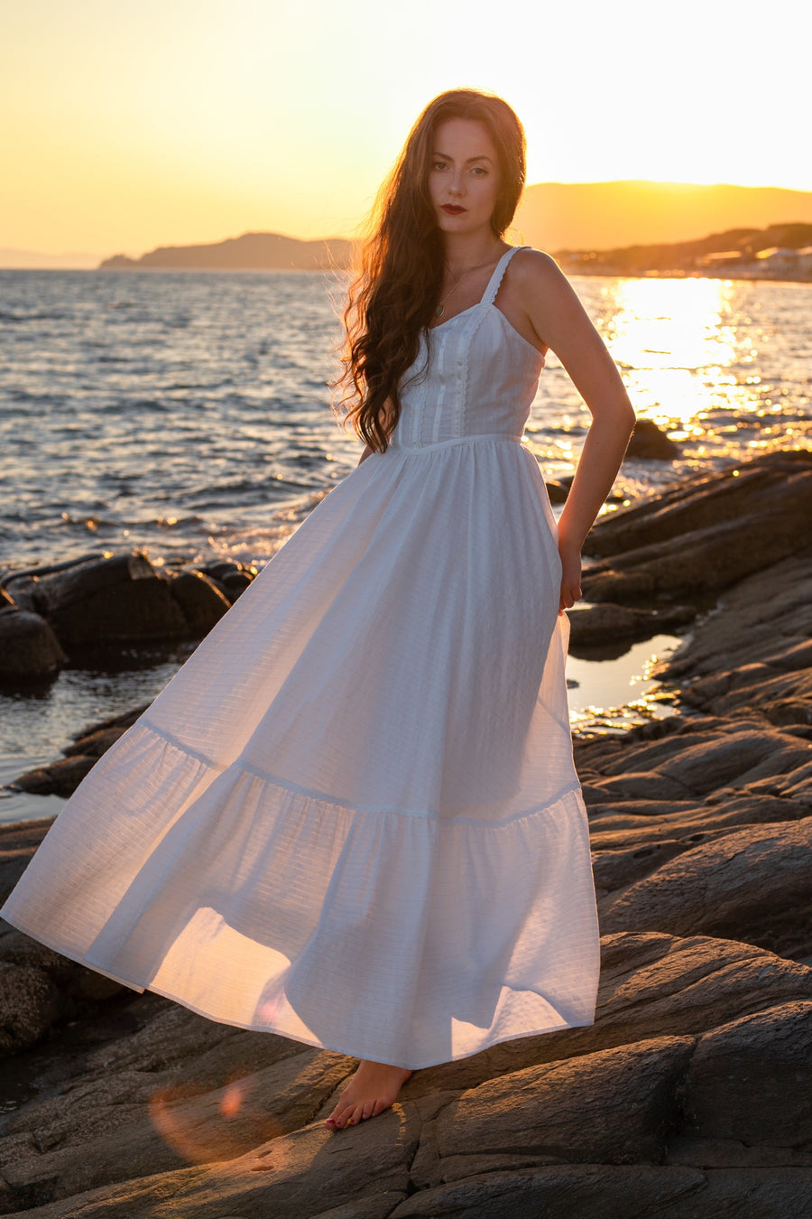 Aphrodite white cotton prairie maxi dress with thin straps, standing on rocks by the sea at sunset