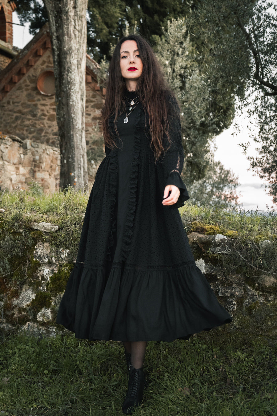 Raven black cotton midi dress with bell sleeves, ruffles and a lace-edged high collar, by an old stone chapel