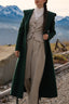Close-up of a sophisticated outfit: beige wool trousers, matching vest, beige suite, lace blouse, and a long green wool coat in an alpine mountain setting; timeless elegance and refined quiet luxury style.