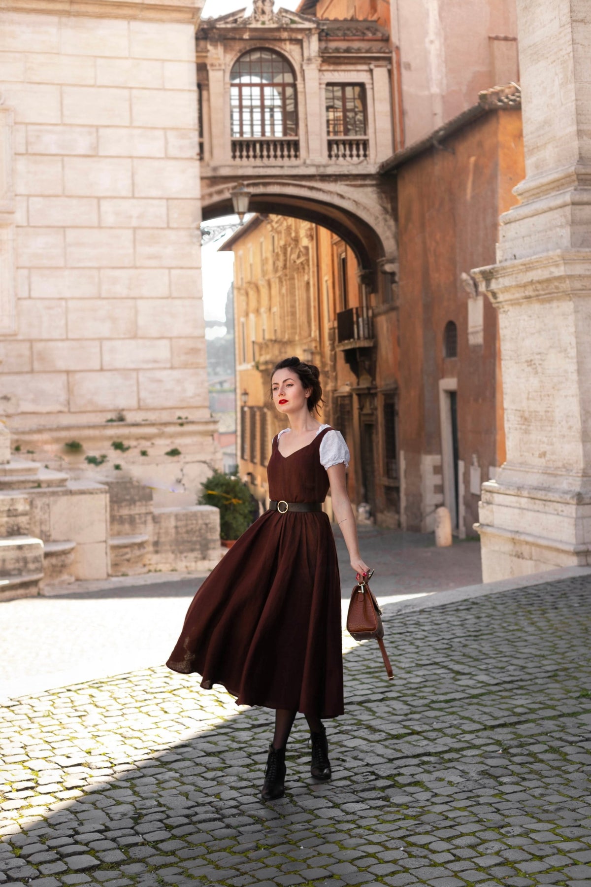 Andalusia dark brown linen midi dress worn over a white puff-sleeve blouse, walking under an arch in Rome