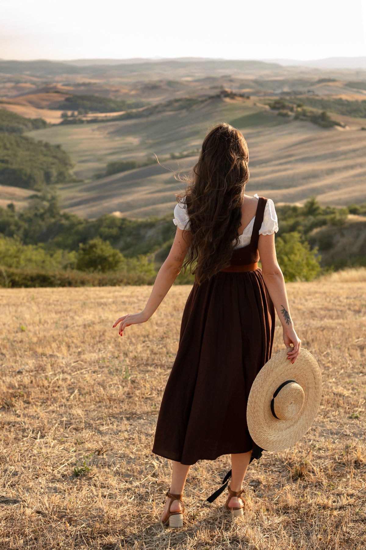 Back view of the Andalusia dark brown linen dress over a white blouse, with a straw hat, overlooking Tuscan hills