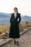 Stylish woman in a belted green wool coat and beige wool trousers standing in a serene mountain landscape; quiet luxury fashion inspired by alpine calm and timeless elegance.
