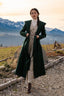 Elegant woman in a deep green wool coat walking along a gravel path with snow-covered alpine mountains in the background; quiet luxury, timeless elegance, wool coat styling in natural landscape.