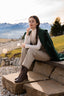 Detail shot of brown lace-up boots, beige wool trousers and a draped green wool coat on stone steps outdoors with alpine scenery; effortless quiet luxury and countryside elegance.